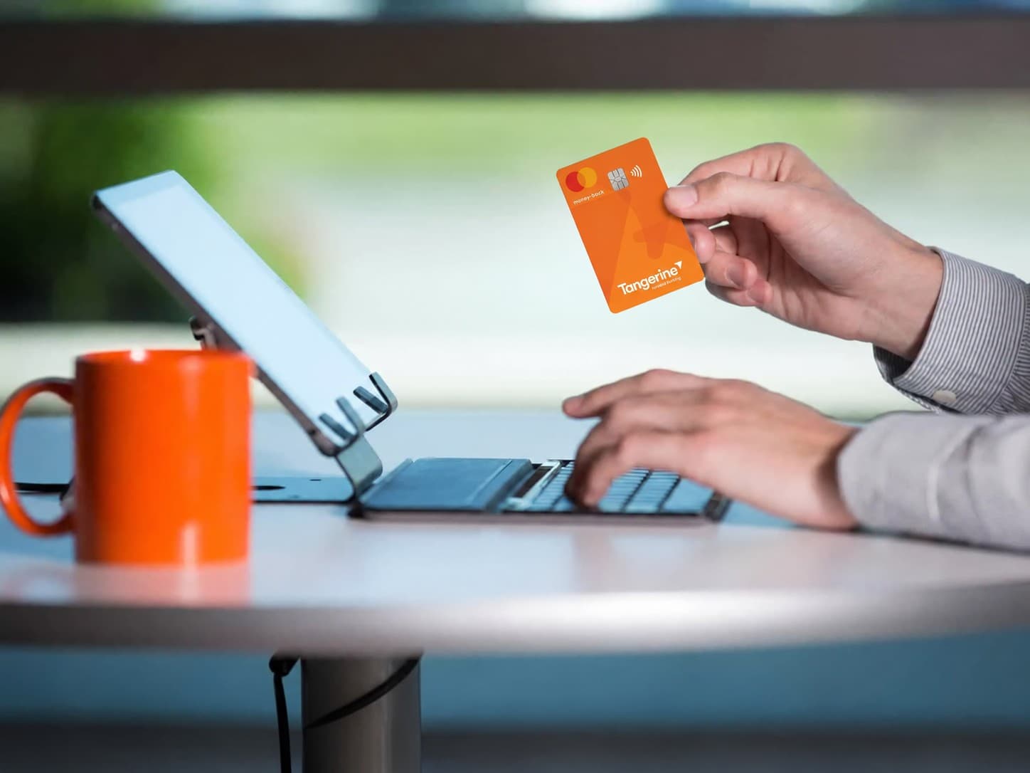 A hand holding a Tangerine Money-Back Mastercard above a laptop keyboard